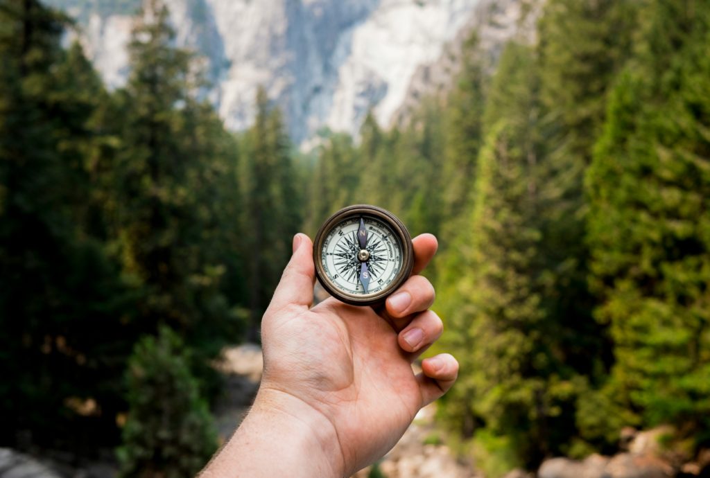 hand holds a compass pointing north in front of a forest of trees